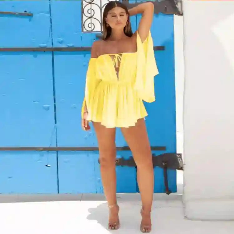 Woman wearing a bright yellow off-shoulder mini dress with wide sleeves and high waist, standing in front of a bold blue door on a sunny day.